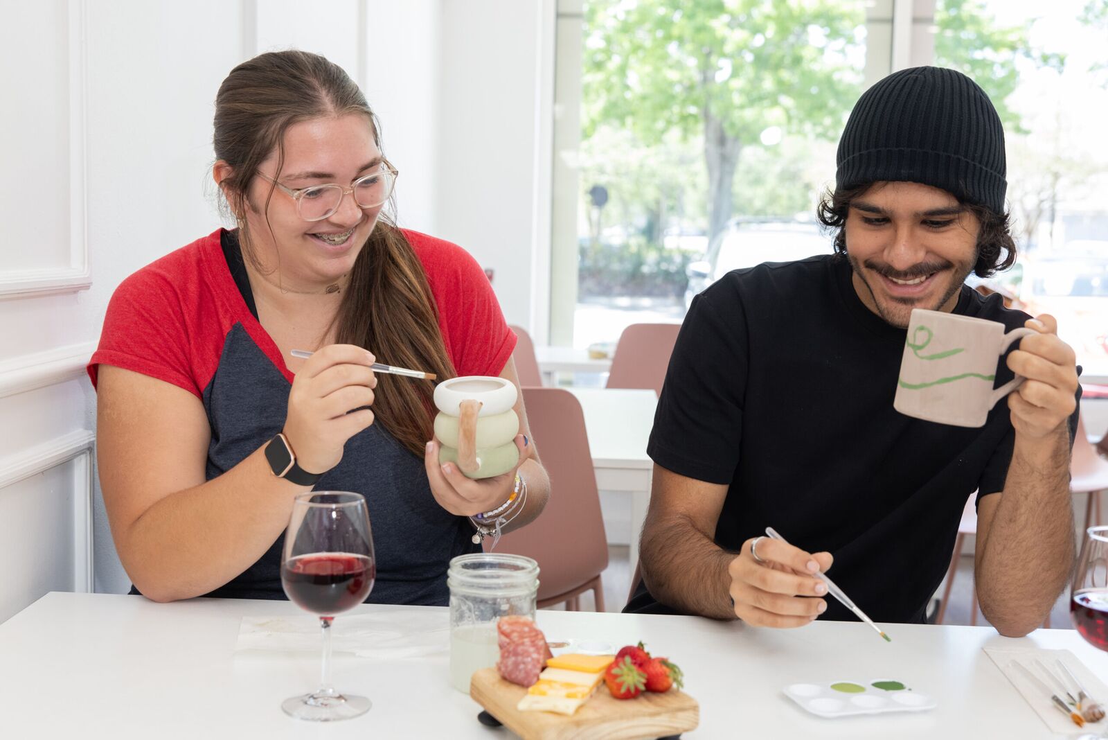 Two friends smiling as they paint mugs, with wine and a cheese board