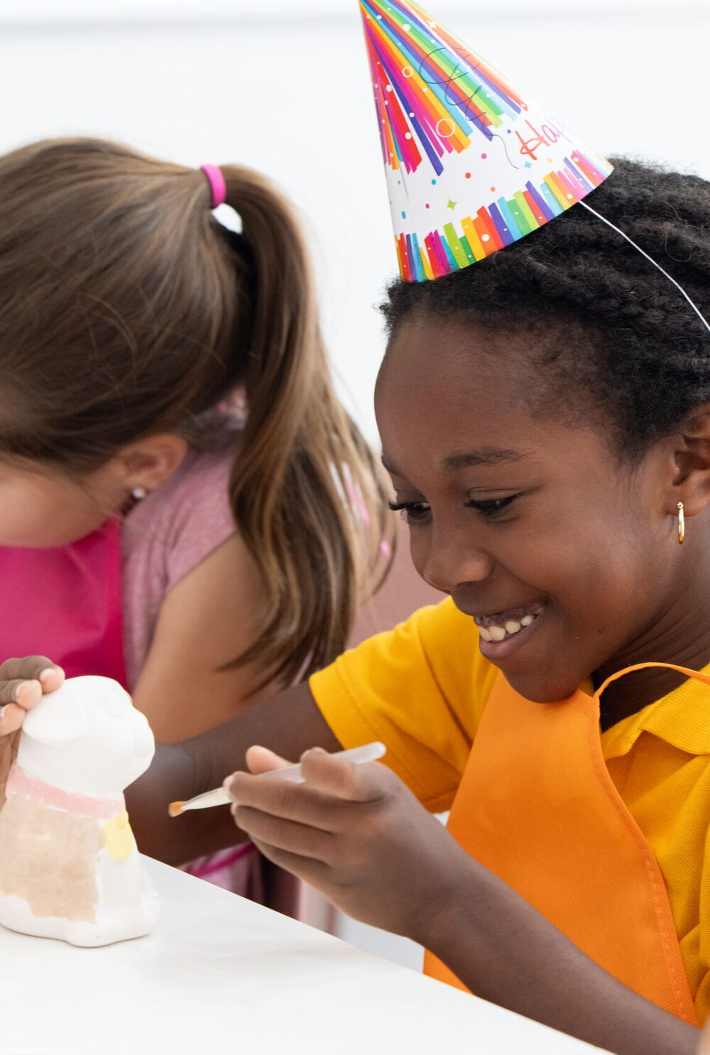 A girl in a party hat smiling as she paints a pottery figure at a birthday party