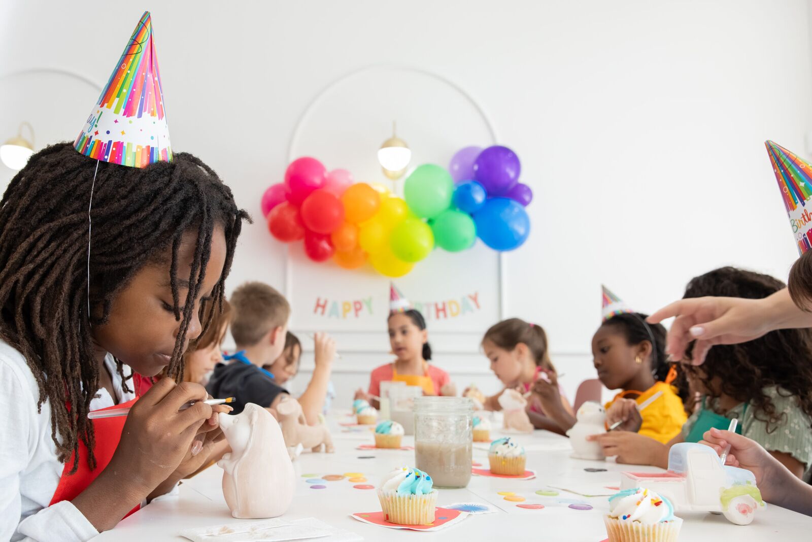 Kids in party hats painting pottery at a long table under rainbow balloons