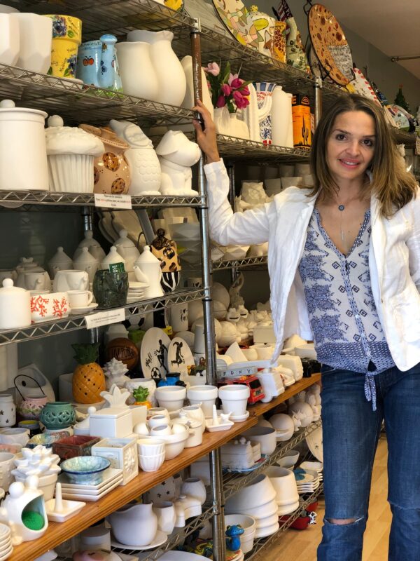 Agi Shah, Owner of Amaze In Pottery Standing by shelves of pottery