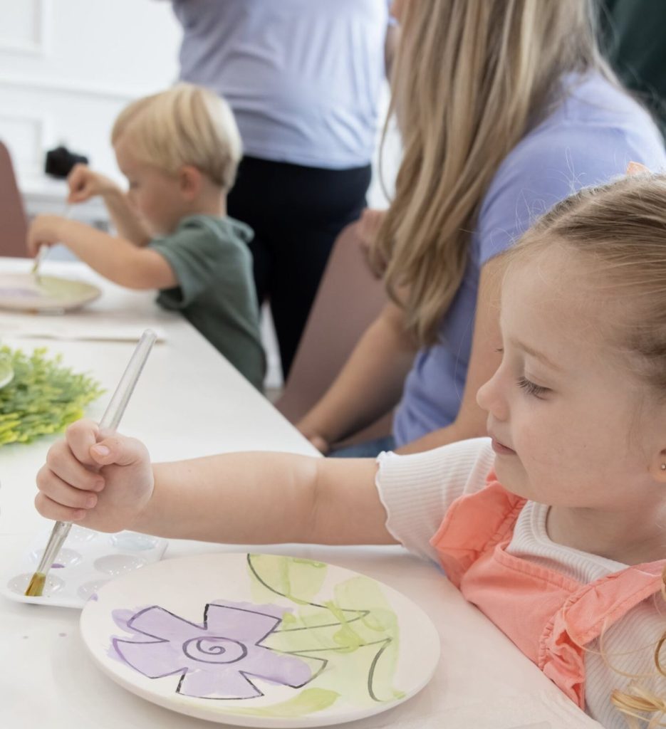 A little girl painting a purple flower plate at the studio, with a parent beside her and another child painting in the background