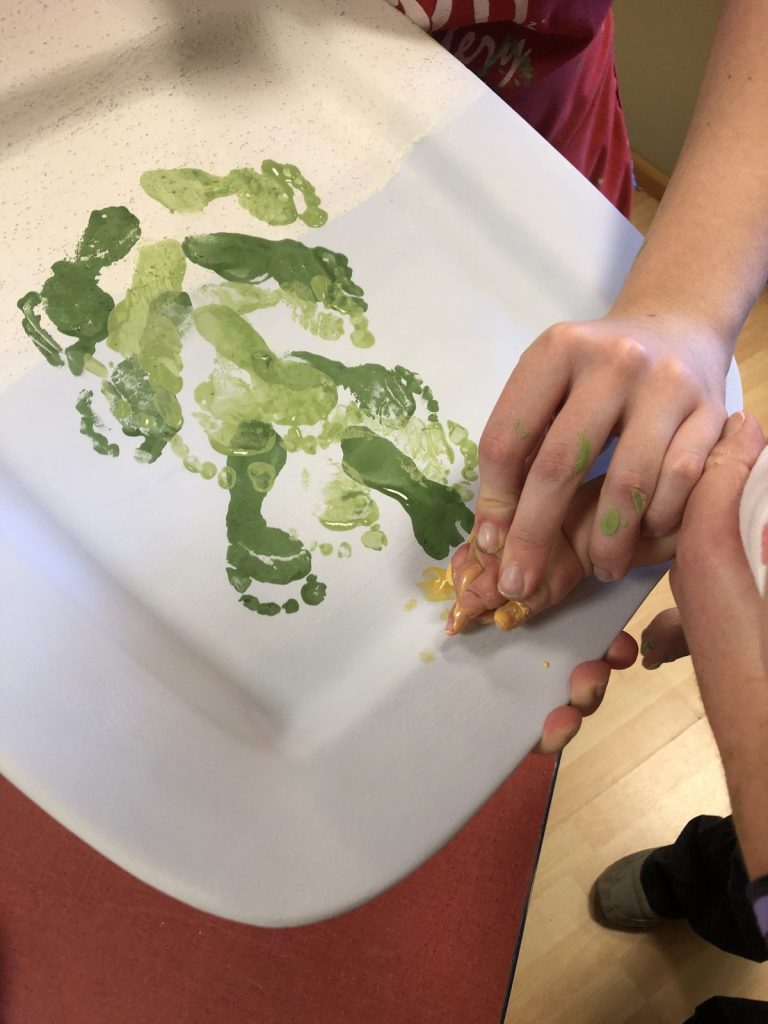 Child's green painted feet being pressed onto a white platter to make a holiday keepsake