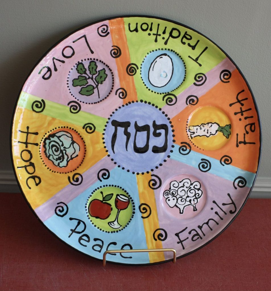 Colorful hand-painted Passover seder plate with Hebrew lettering, symbolic foods and words like Love and Hope