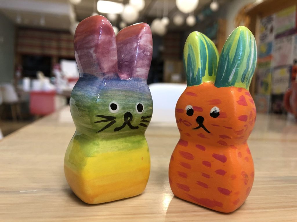 Two hand-painted ceramic bunnies, one in rainbow stripes and one orange with green ears