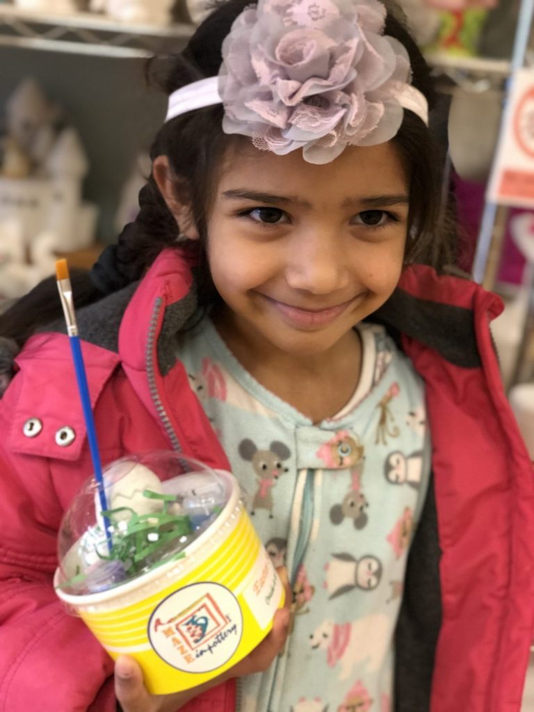 Smiling girl holding a paint-at-home kit cup with a brush in the studio