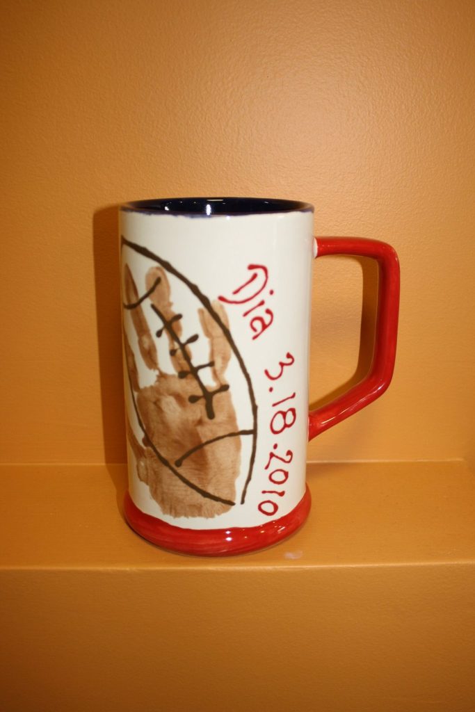 Hand-painted tall mug with a brown handprint football and red handle and base, dated 2010