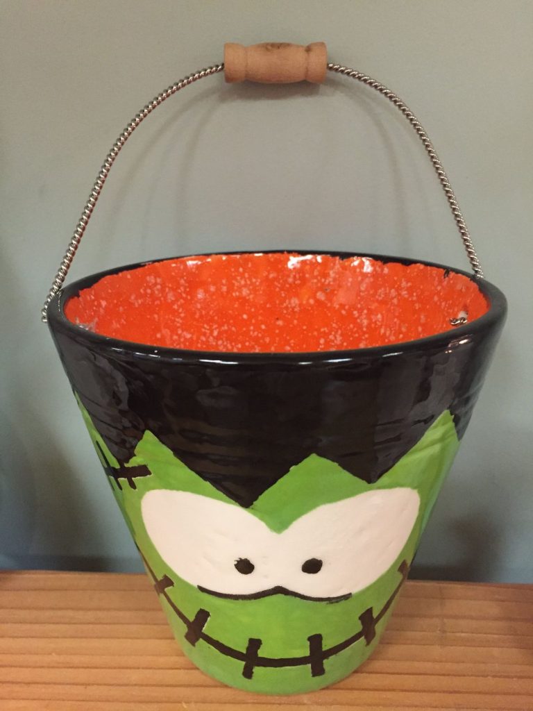 Hand-painted ceramic bucket with a green Frankenstein face, orange interior and wire handle