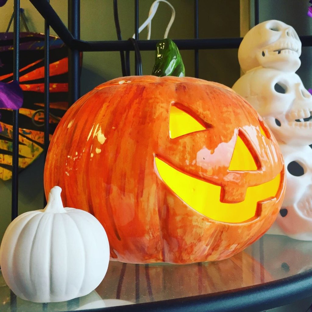Hand-painted orange jack-o'-lantern glowing from inside beside a white pumpkin and stacked skulls