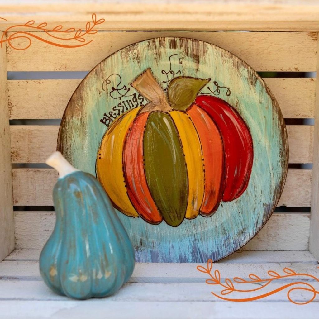 Hand-painted round platter of a rainbow-striped pumpkin on a weathered teal background beside a teal gourd