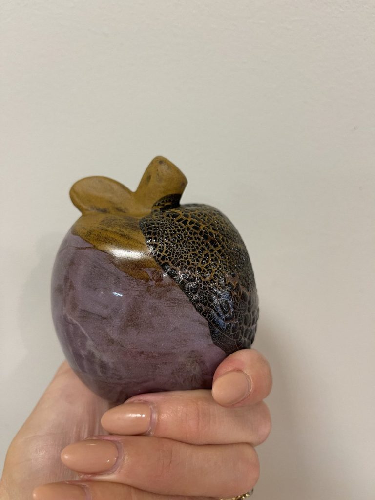 Hand-painted apple in purple and black crackle glaze with a bronze stem, held in hand
