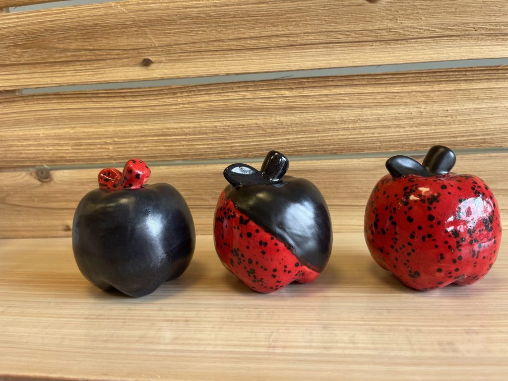 Three hand-painted apples in glossy black and speckled red glazes on a wooden shelf