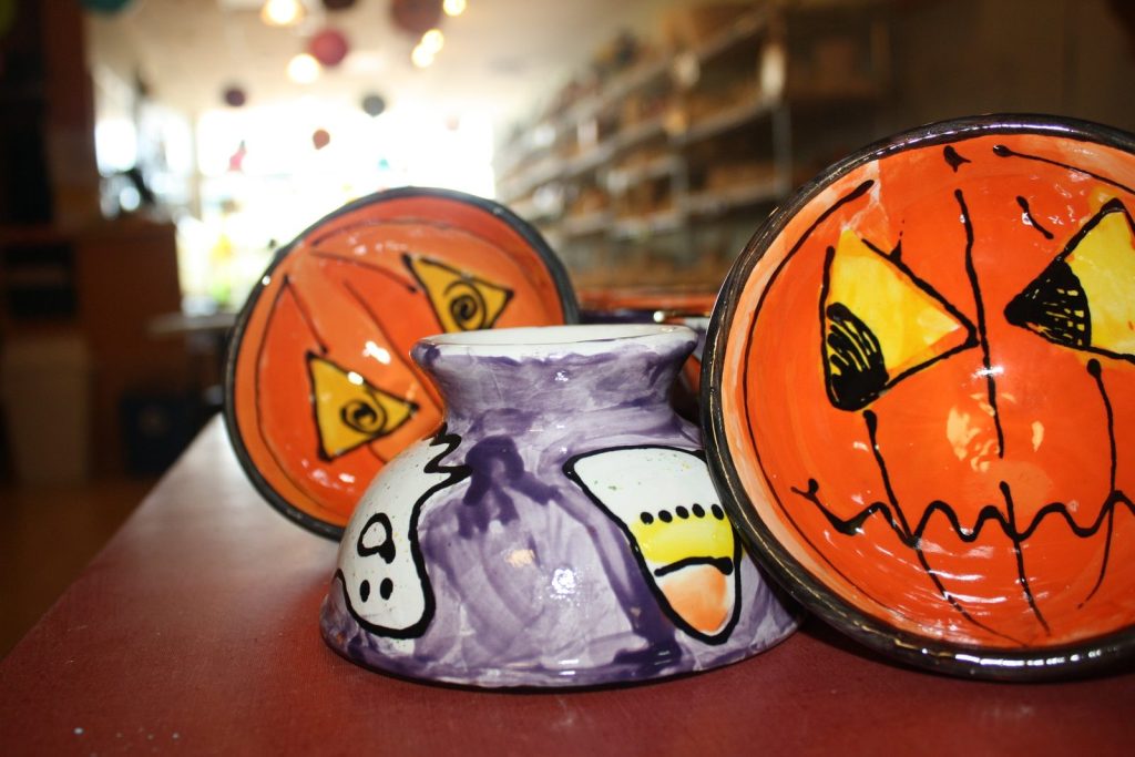 Hand-painted orange jack-o'-lantern bowls beside a purple piece with a ghost and candy corn