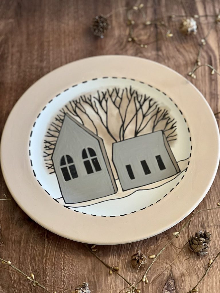 Hand-painted plate of two gray houses under bare winter trees with a dashed border