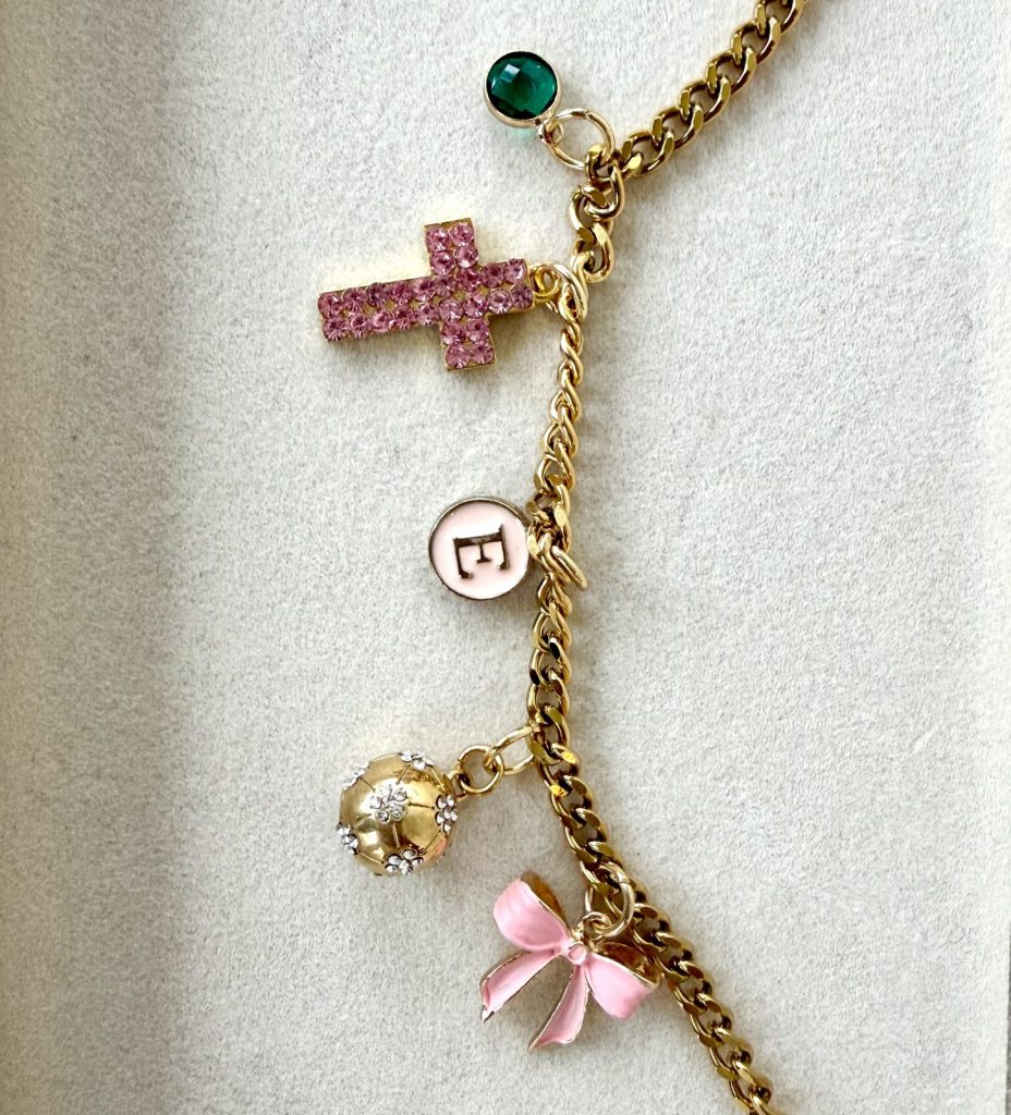 Gold charm necklace with an initial E, a pink cross, a bow and a green stone