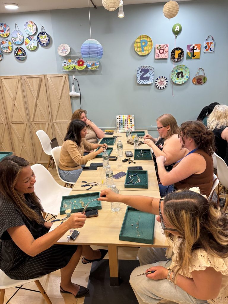 An adult Charm Bar party around a long table at the studio, each guest with a tray of charms