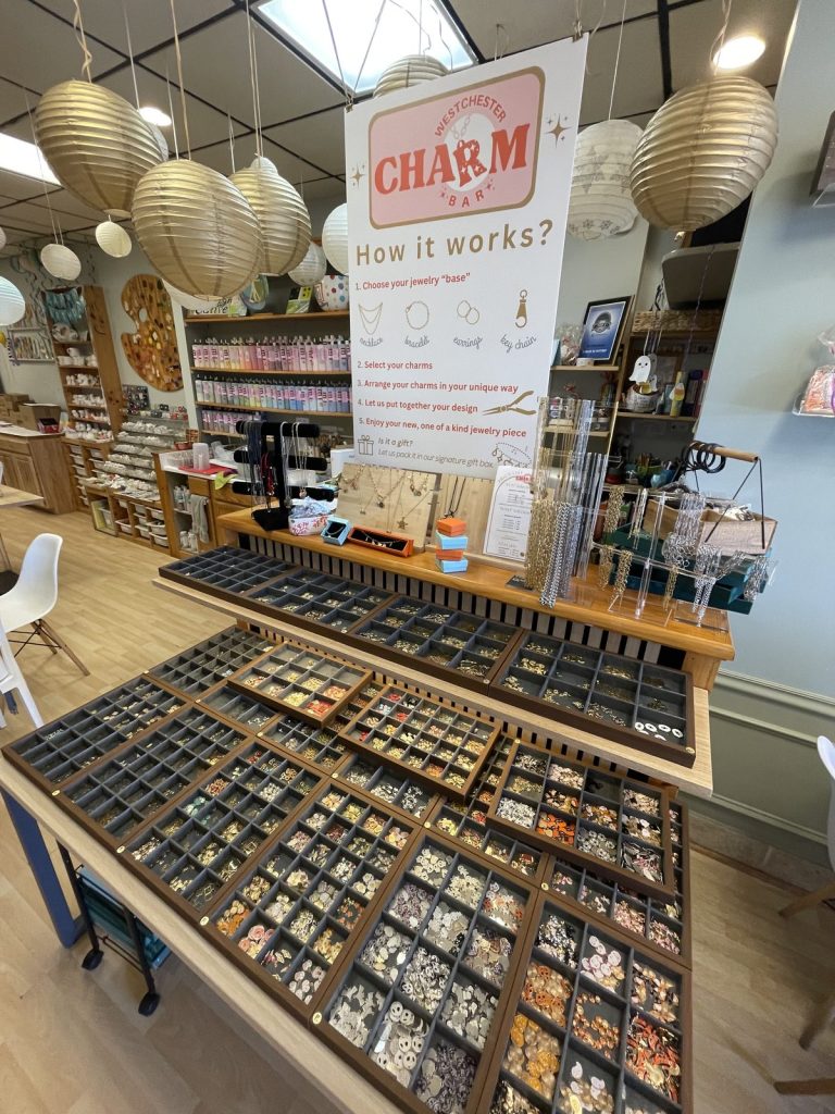 The Westchester Charm Bar counter at our Briarcliff Manor studio, with trays of charms and a How it works sign