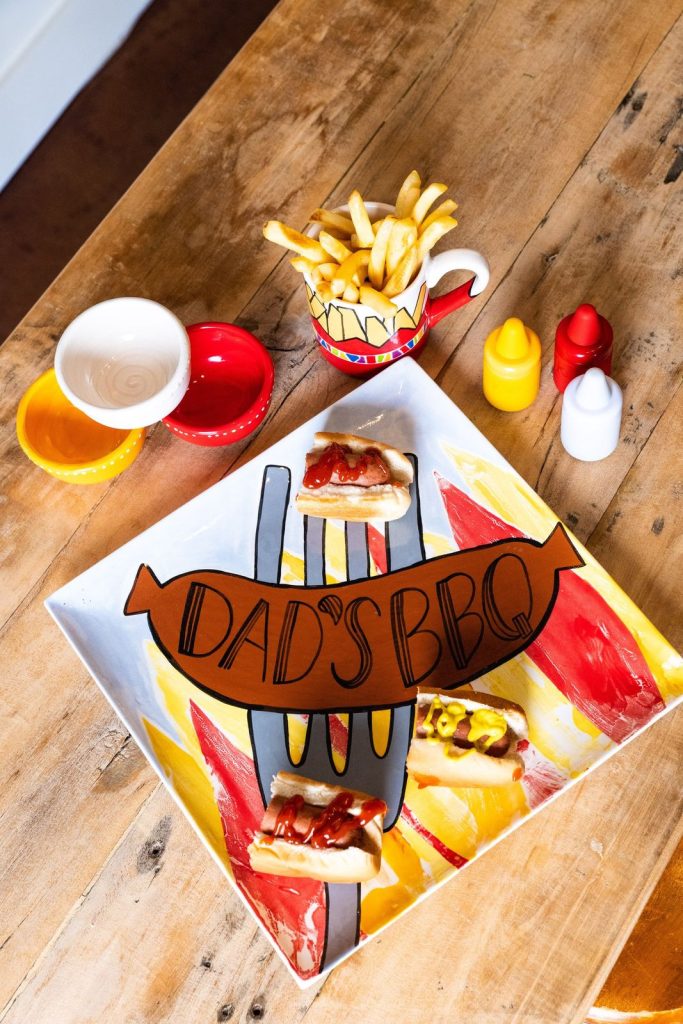 Square Dad's BBQ plate with painted hot dog and fork, served with hot dogs and fries