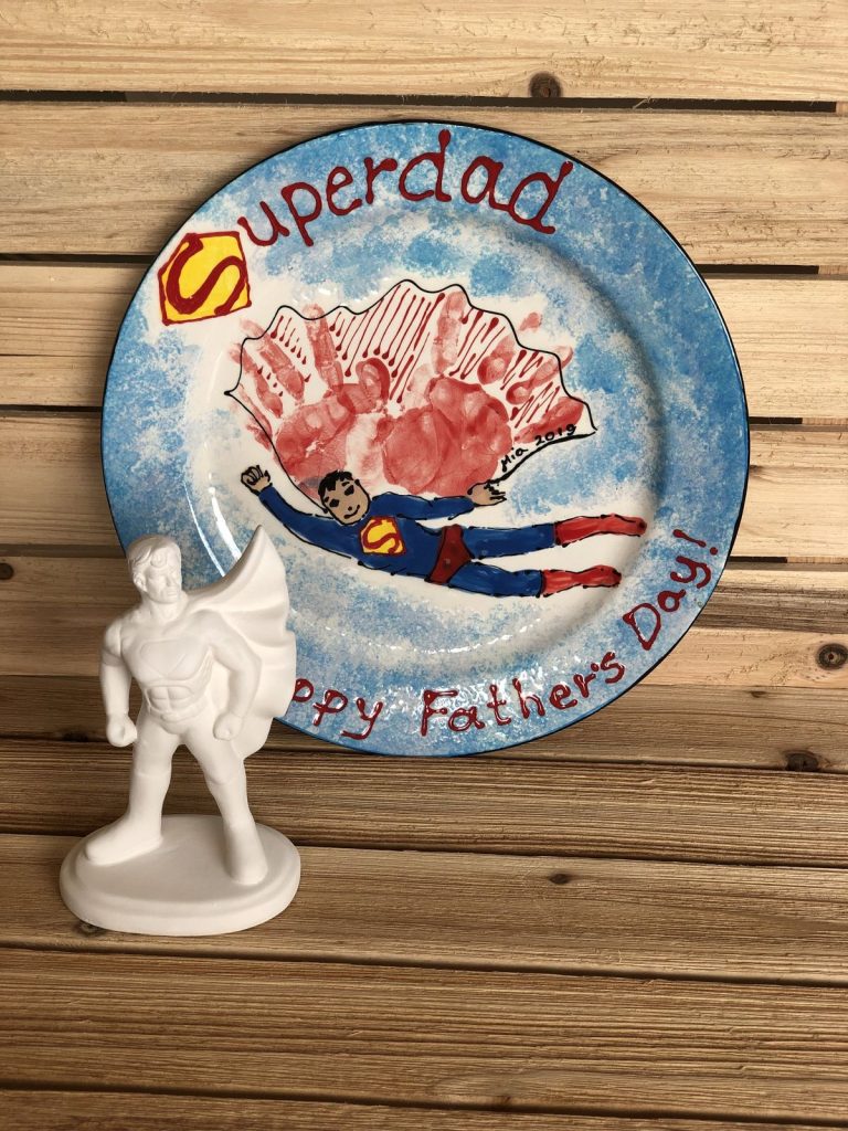 Round Superdad Father's Day plate with red handprint cape and flying superhero, beside figurine