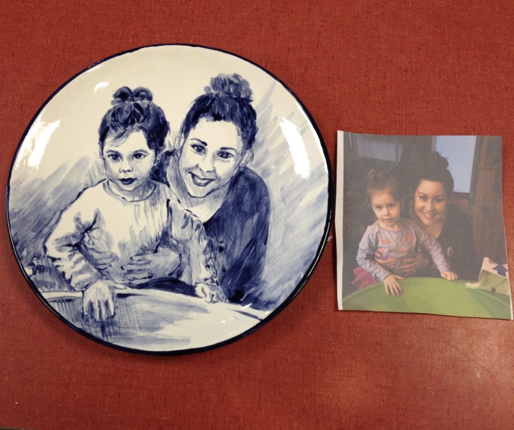 Hand-painted blue and white round plate portrait of a mother and daughter beside the reference photo