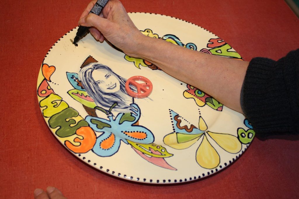 Artist outlining a hand-painted round plate with a girl's portrait, peace signs and butterflies