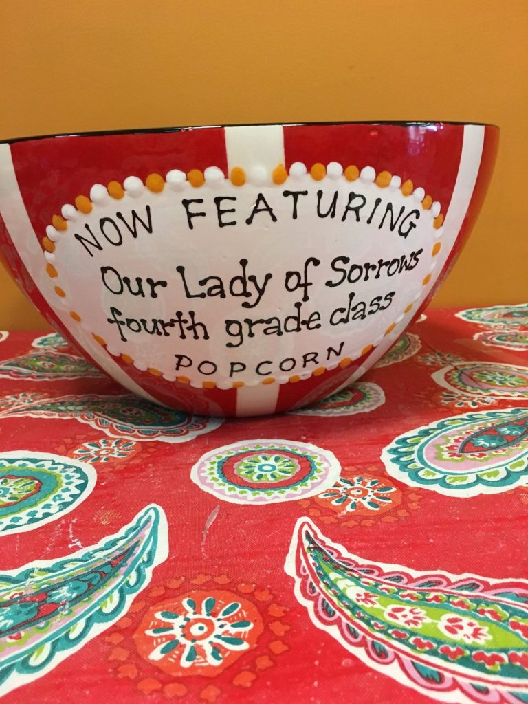 Hand-painted red-striped popcorn bowl reading "Now Featuring Our Lady of Sorrows fourth grade class"
