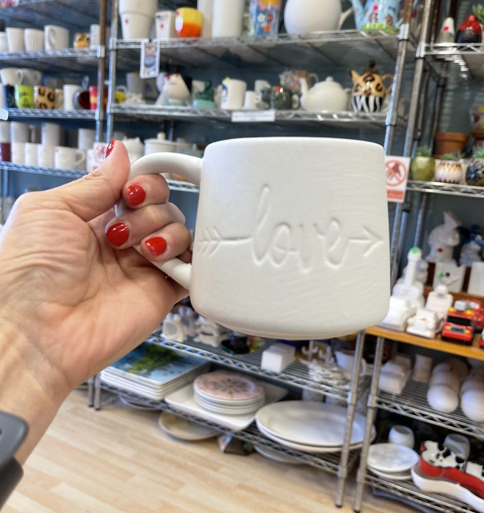Unpainted white bisque mug with raised love lettering and an arrow, held up in the studio